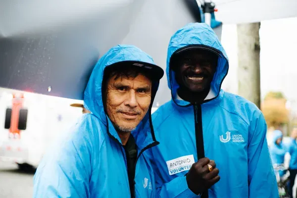 Two men in blue raincoats, helping welcome guests.